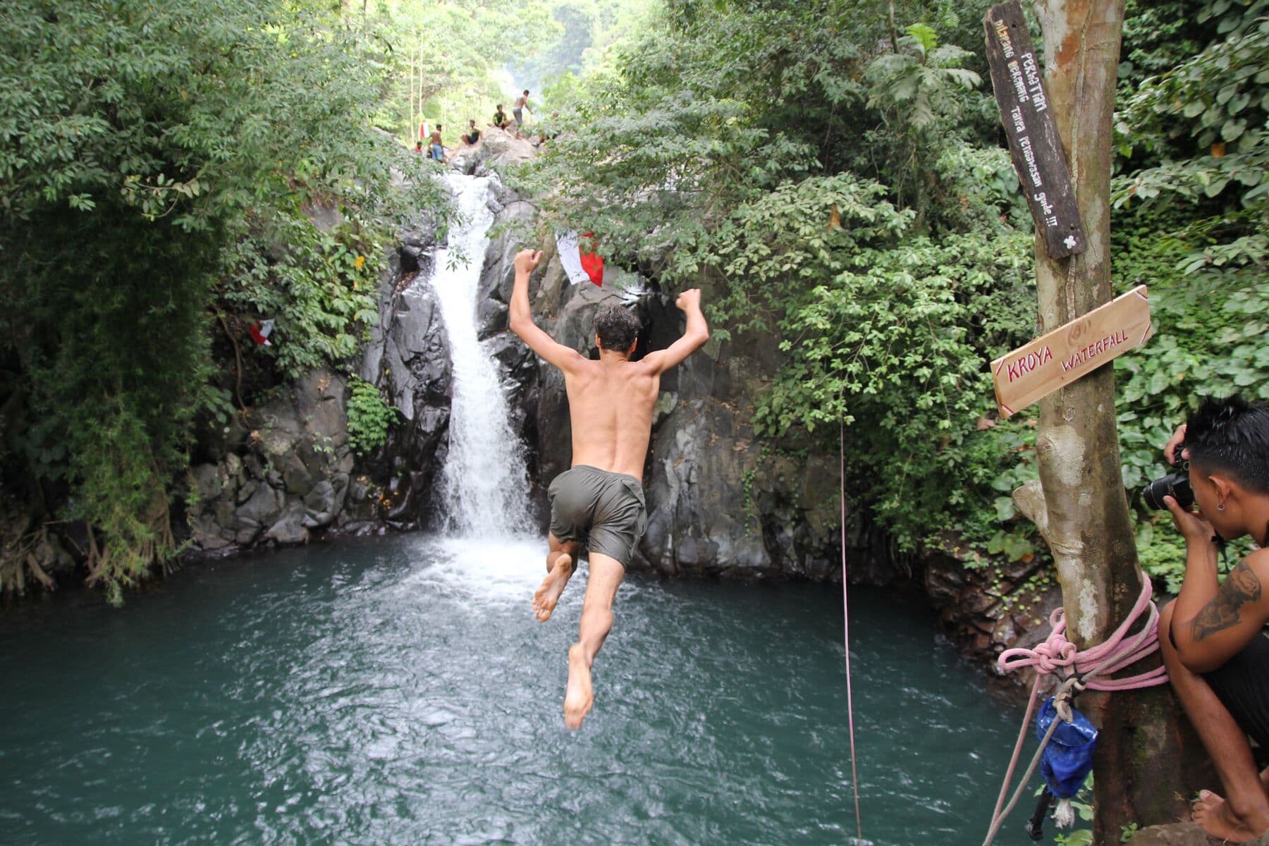A man jumping into a pool below a jungle waterfall, with a photographer at the side
