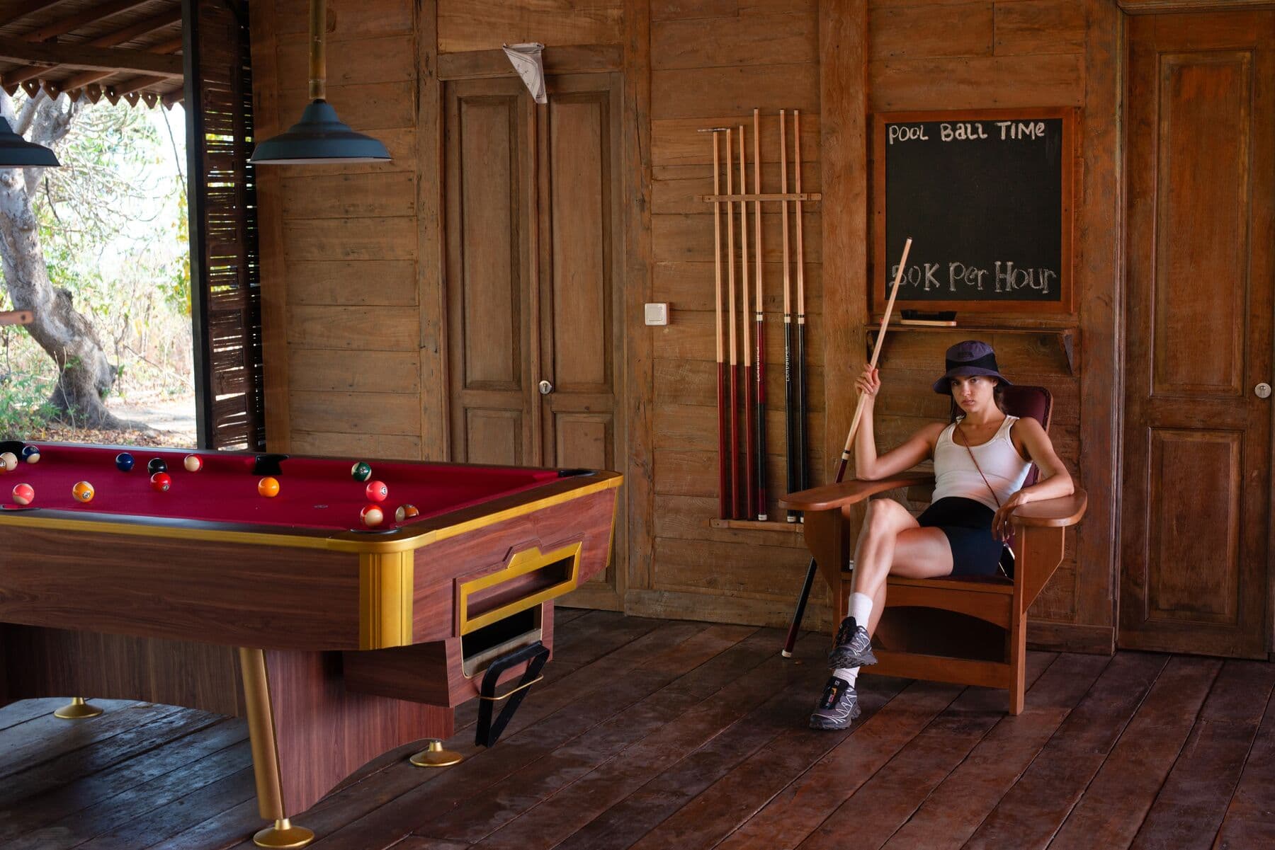 A woman holding a pool cue sits in a chair beside a pool table in a wooden room