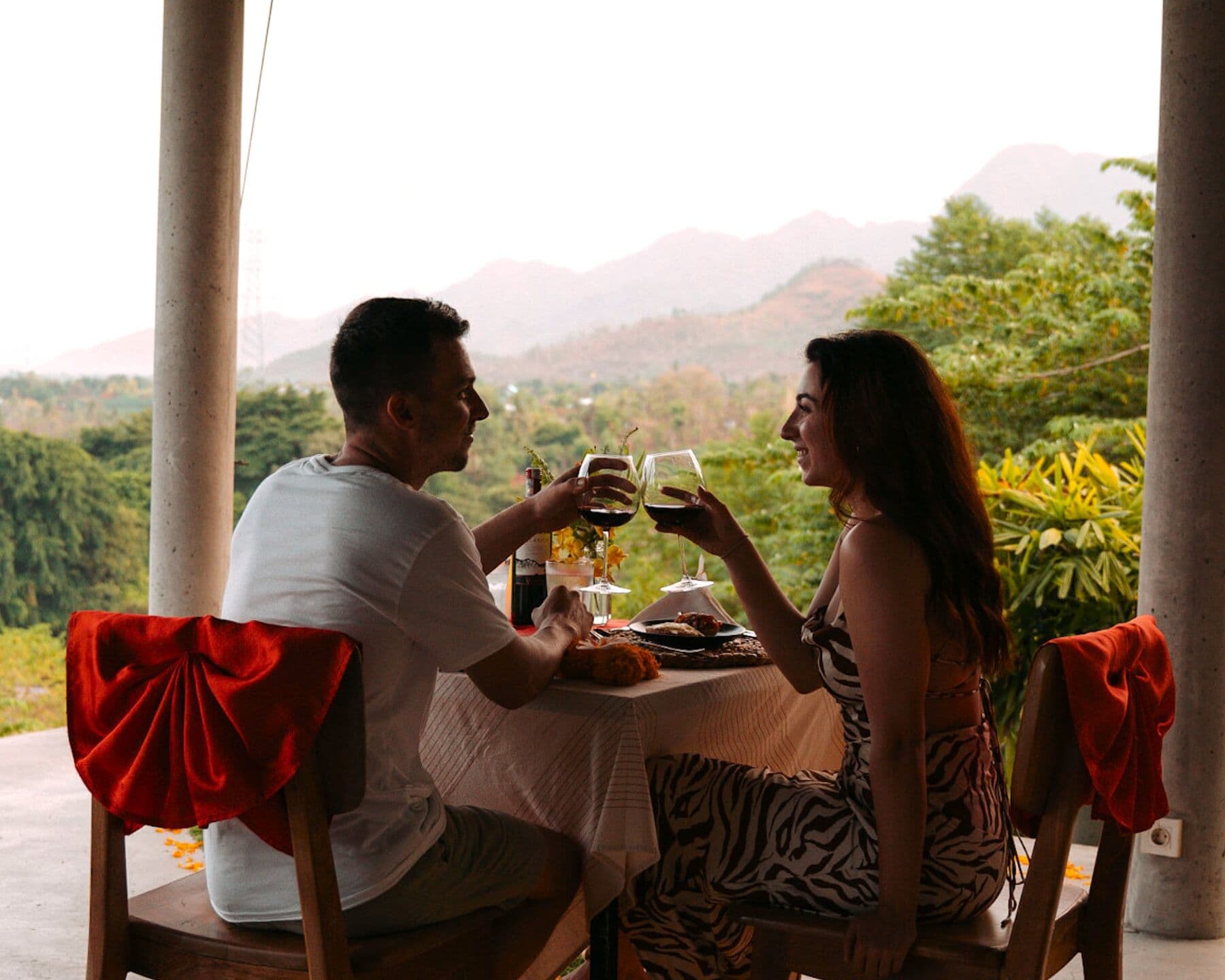 A couple toasting with red wine at a dinner table, with green hills behind