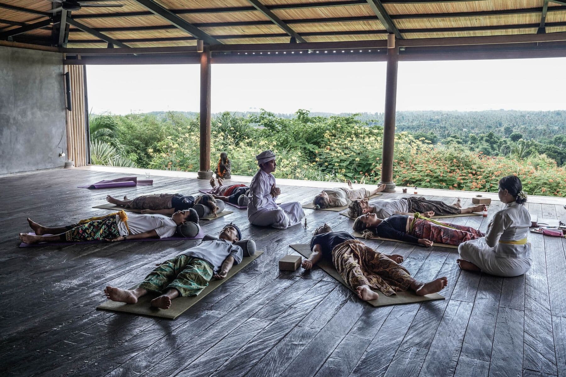 People lying on mats in an open wooden shala during a ceremony, with a woman in white seated beside them