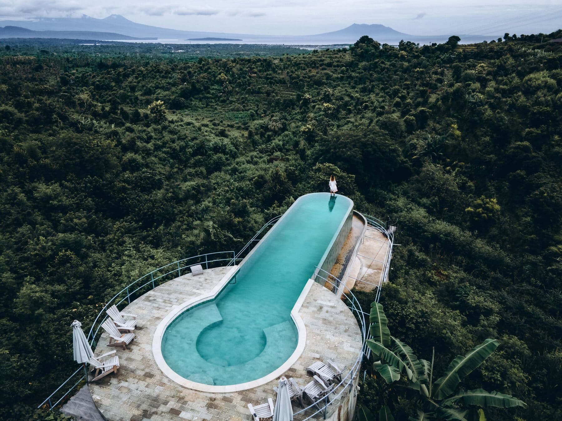 Aerial view of a long infinity pool with sun loungers, surrounded by forest