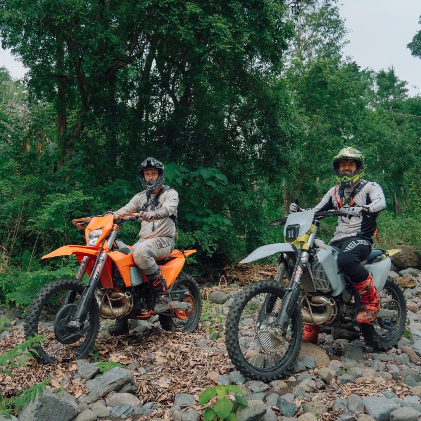 Two riders in helmets sitting on an orange and a white dirt bike on a rocky riverbed in the forest