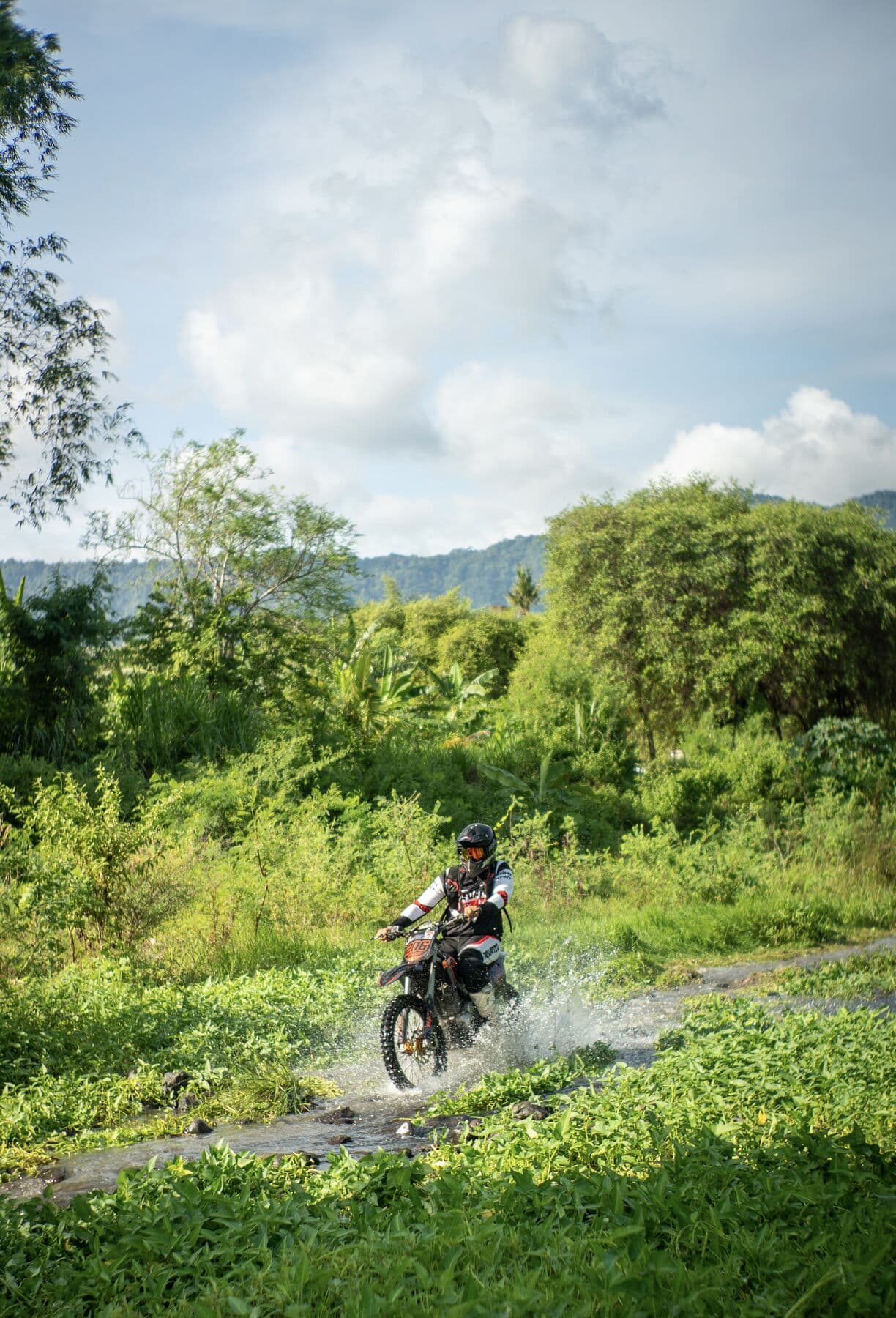 A rider in a helmet and goggles splashing a dirt bike through a stream on a jungle trail