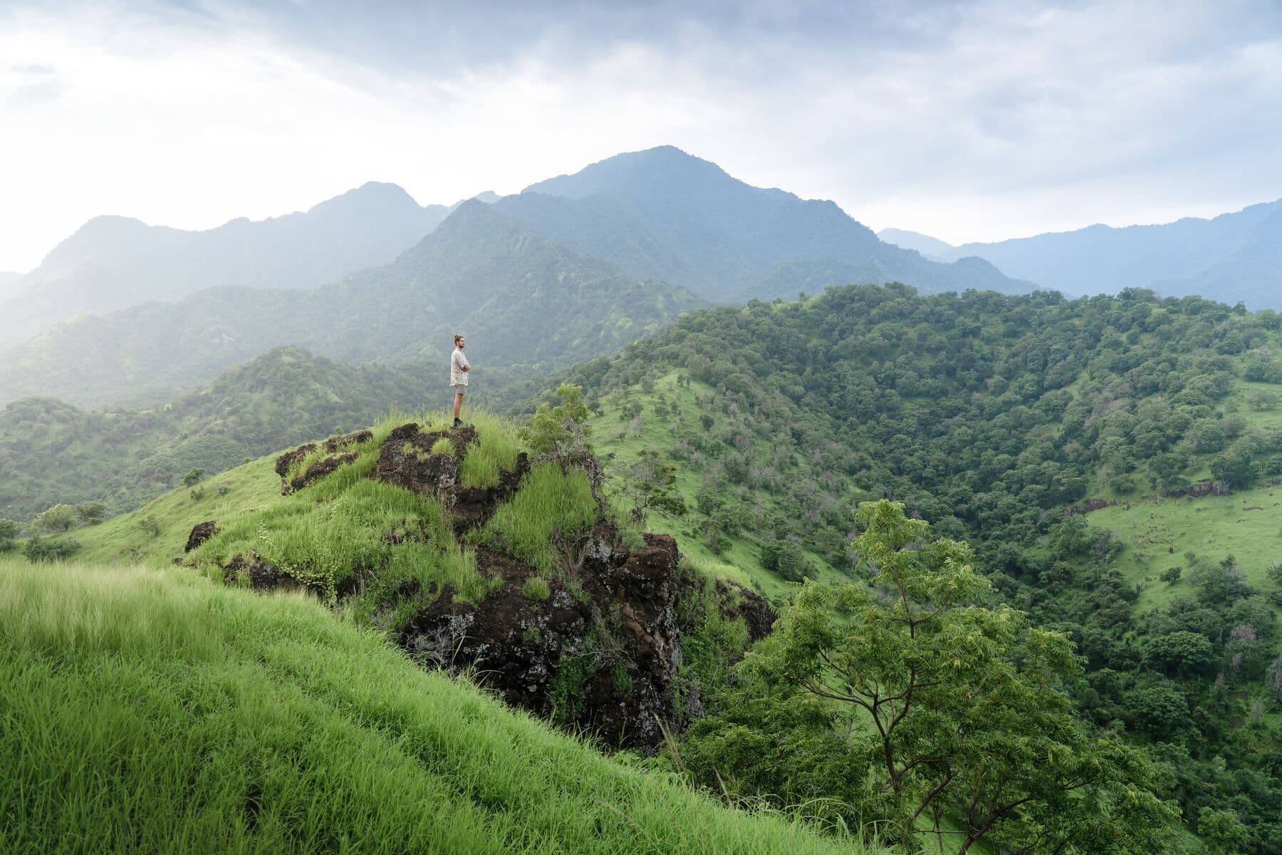 A hiker standing on a rocky green hilltop with forested mountains behind