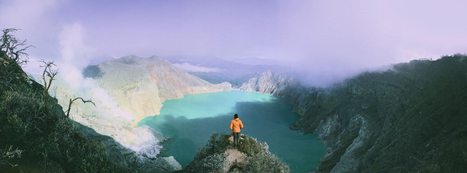 A hiker in an orange jacket standing above the turquoise crater lake of Ijen