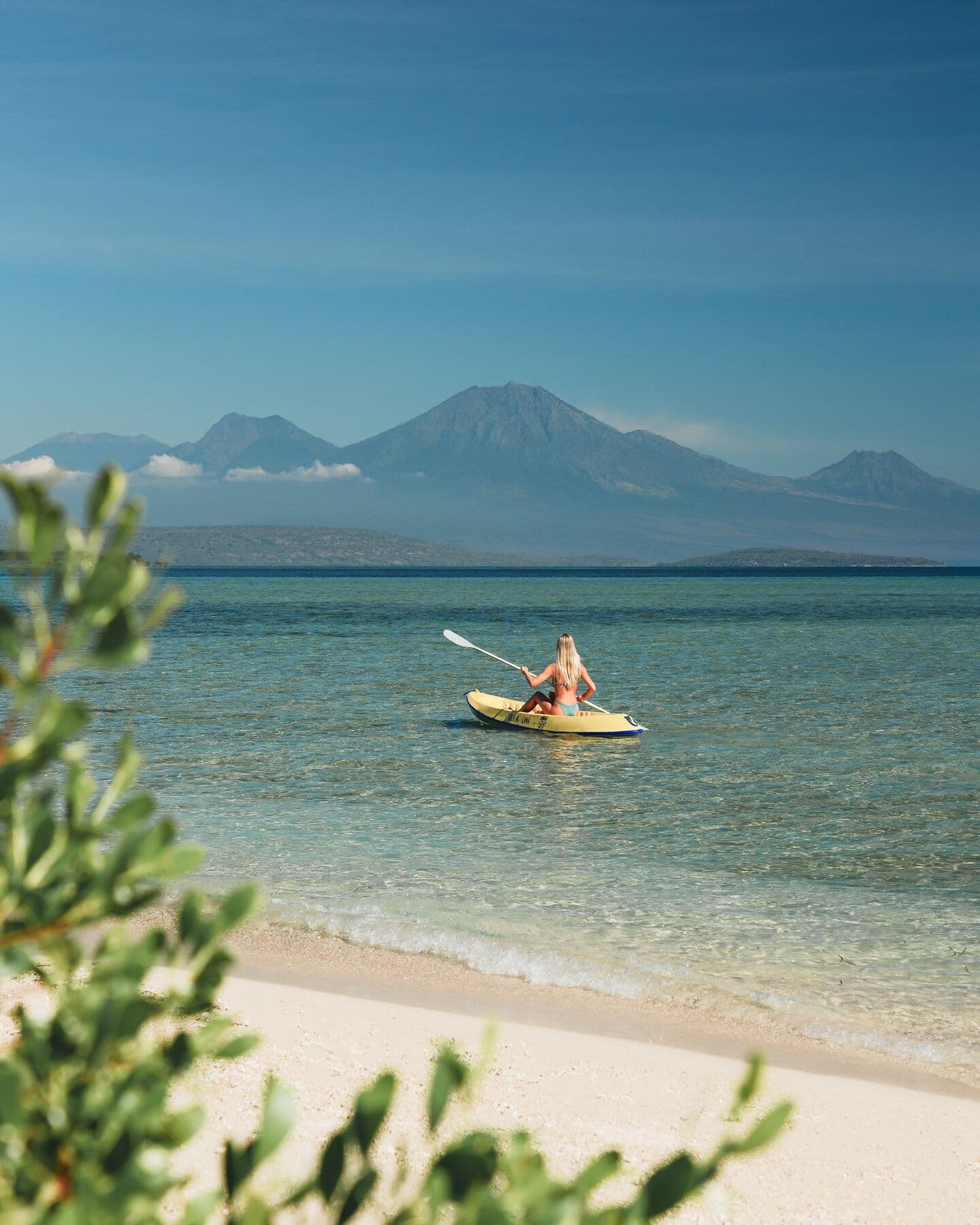 A woman paddling a yellow kayak near the shore, with the volcanoes of Java across the water