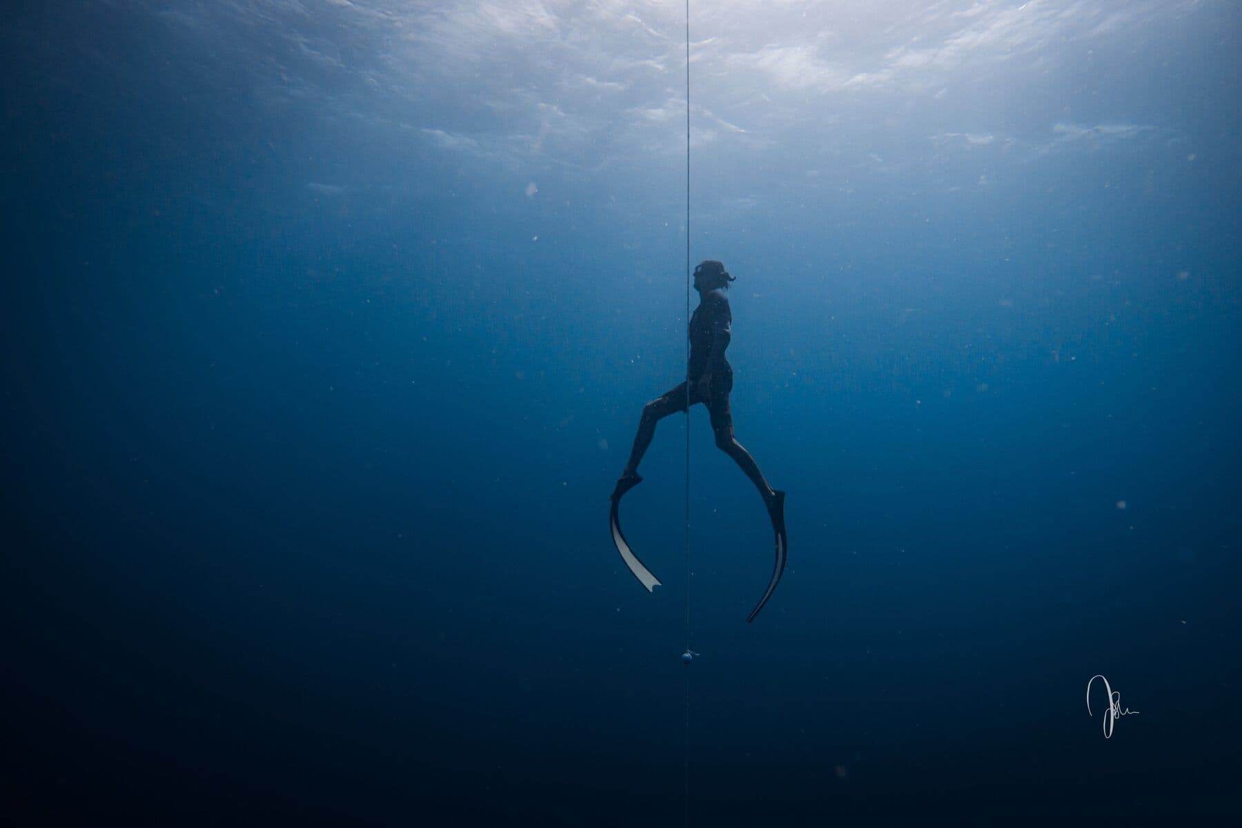 A freediver with long fins ascending beside a vertical line in deep blue water