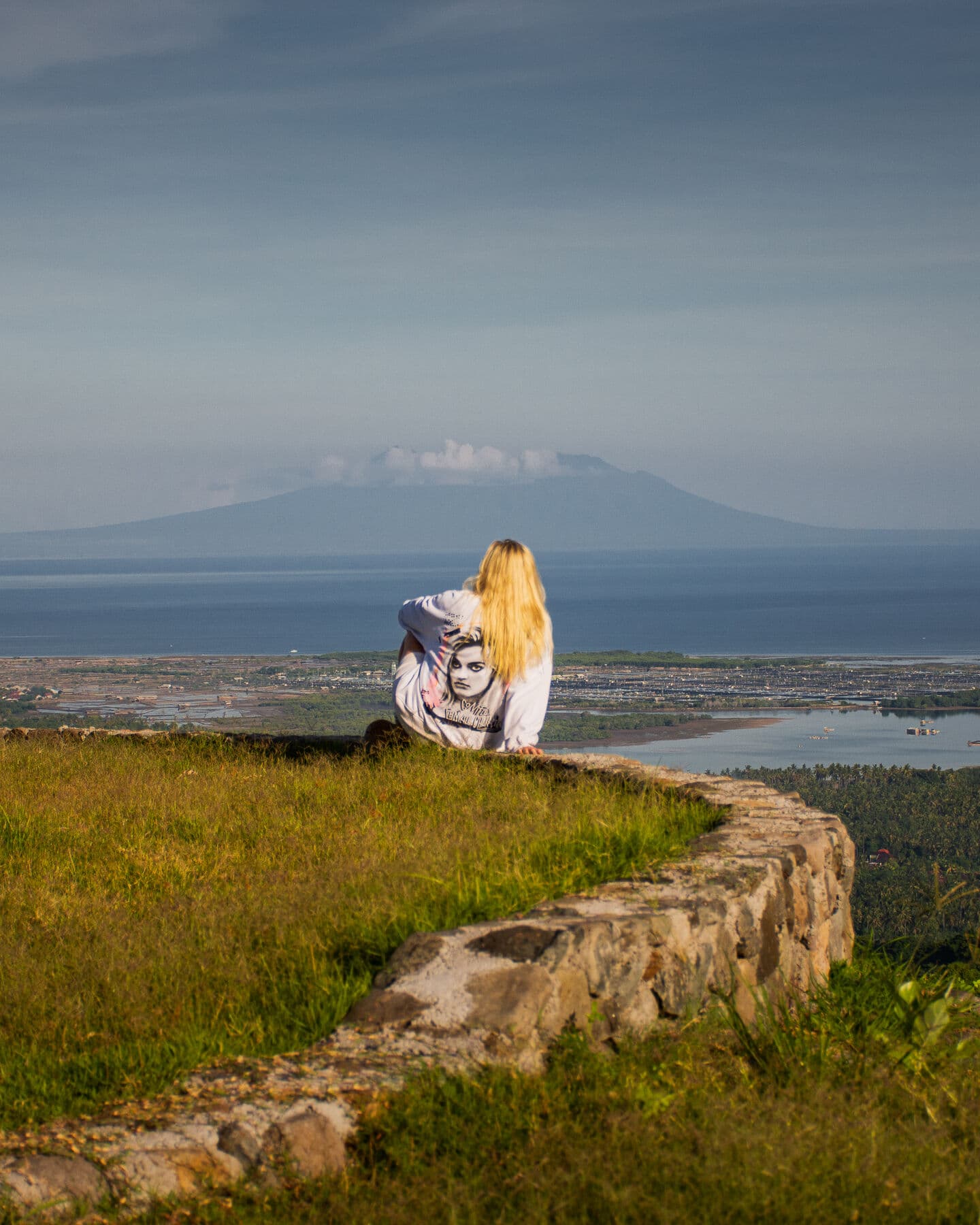 A woman sitting on a stone wall on a grassy hilltop, looking over the coast towards a volcano