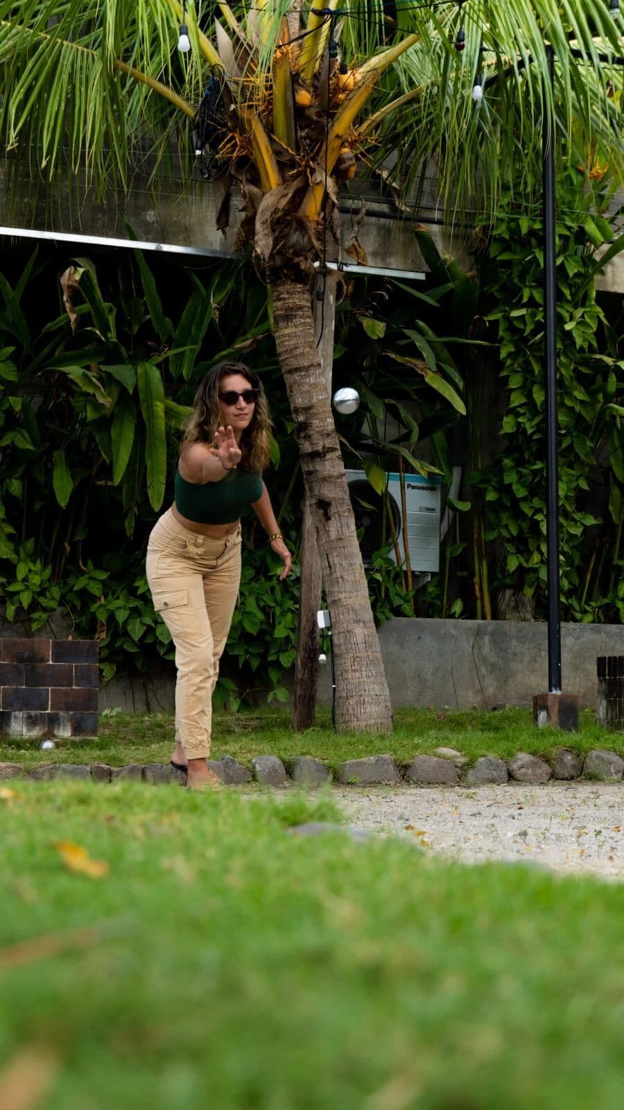 A woman throwing a pétanque ball on a lawn under palm trees