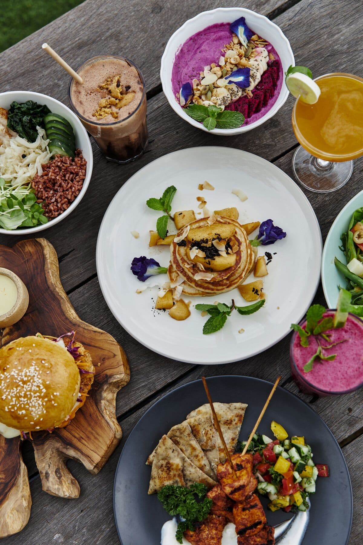 Dishes and drinks seen from above on a wooden table: a burger, pancakes, smoothie bowls, salads and chicken skewers