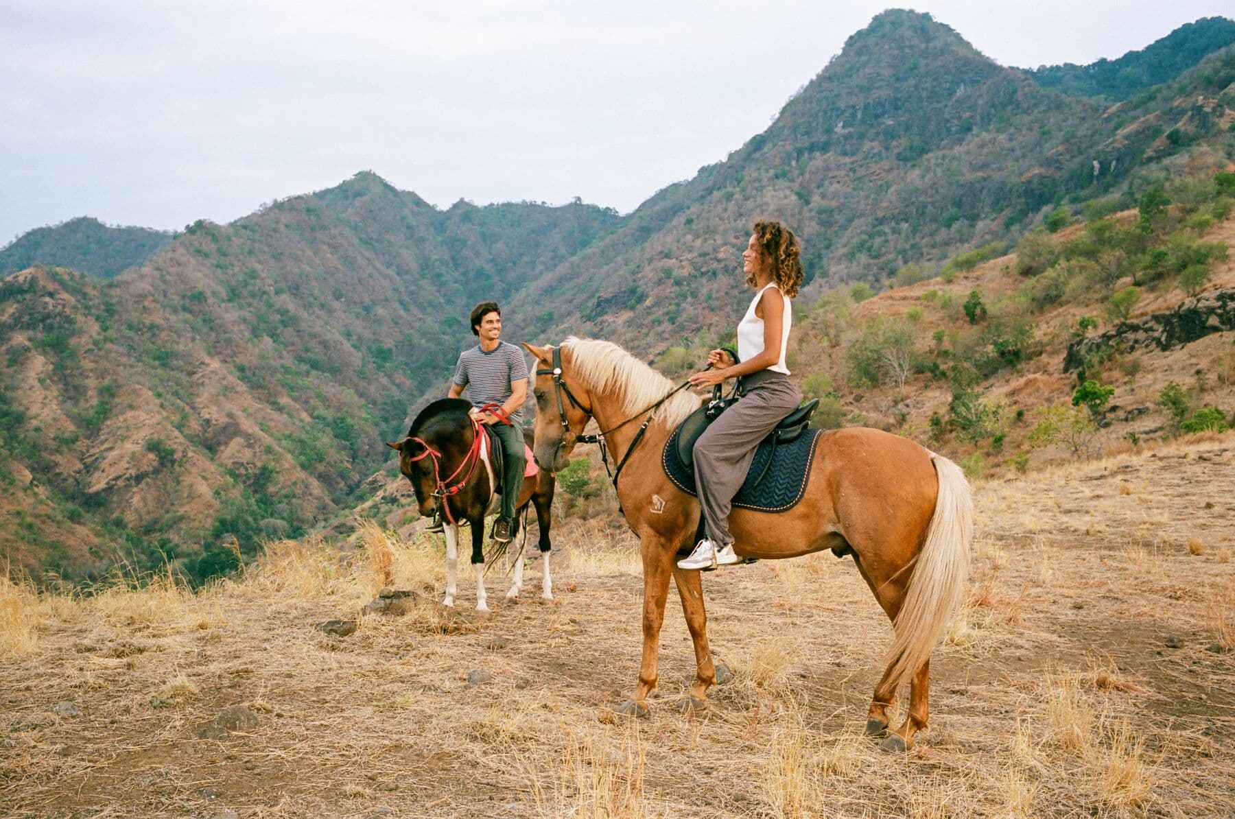 Two riders on horseback on a dry grassy ridge with green mountains behind