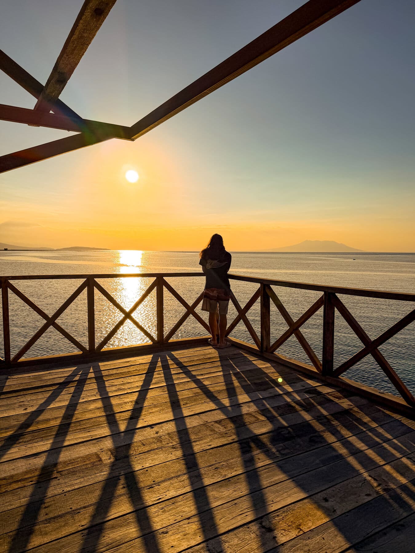 A woman standing at the wooden railing of the outpost deck, looking at the sun setting over the sea