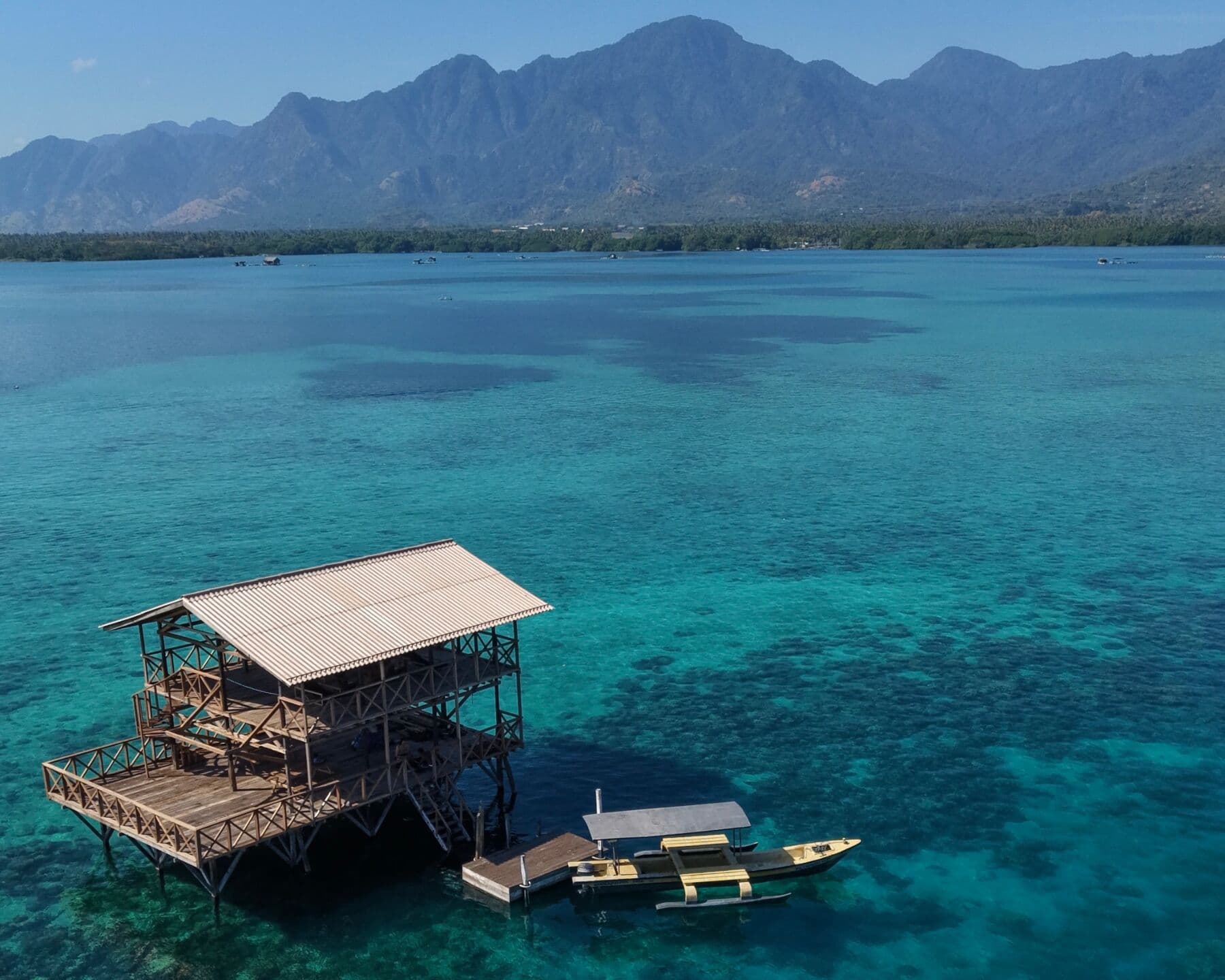 Aerial view of a two-story wooden platform over a reef in turquoise water, with a boat moored beside it and mountains behind