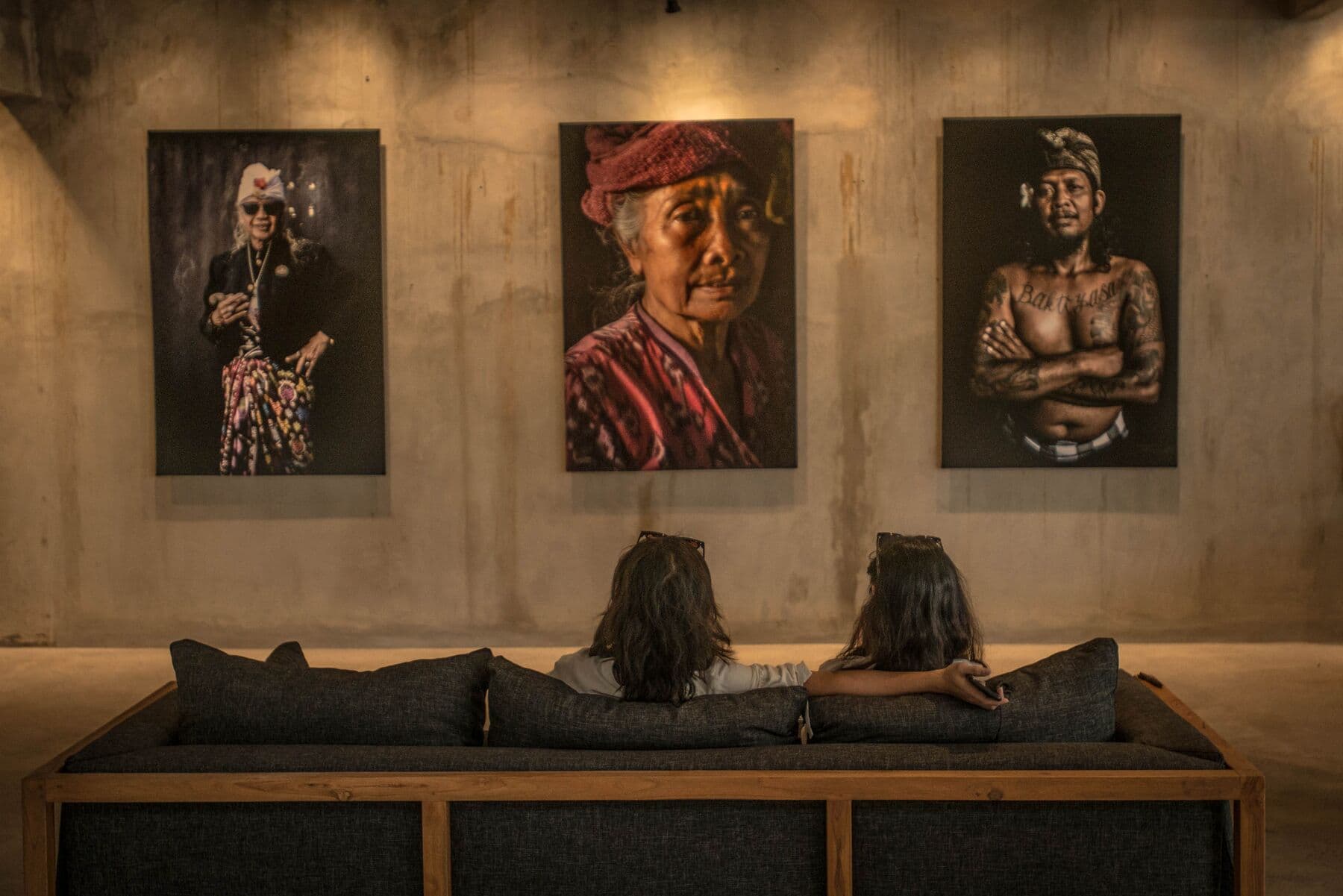 Two people on a sofa looking at three large portrait photographs of Balinese people on a concrete wall