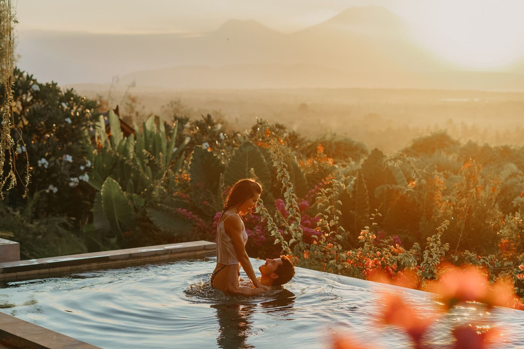A couple embracing in an infinity pool at sunset, with flowers and hills behind