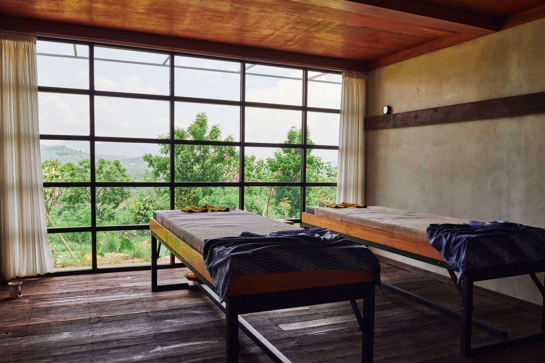Two massage beds in a treatment room with large windows looking onto trees and hills