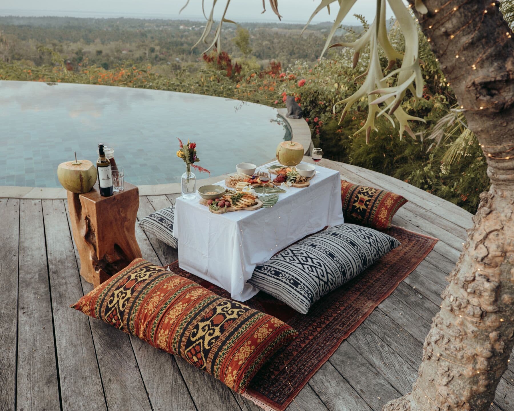 A low table with food, wine and coconuts, set on a rug with cushions beside an infinity pool