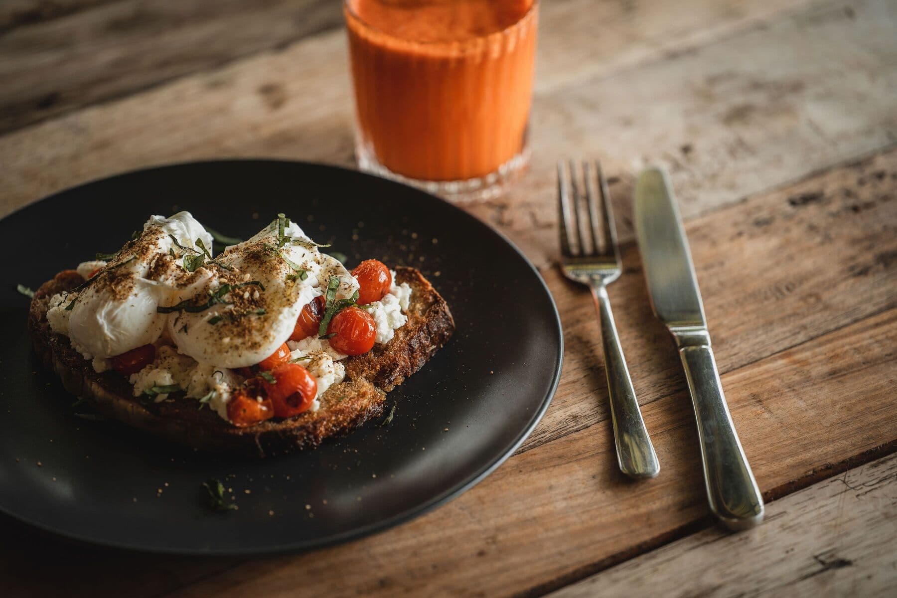 Poached eggs and roasted cherry tomatoes on toast, with a glass of red juice, on a wooden table