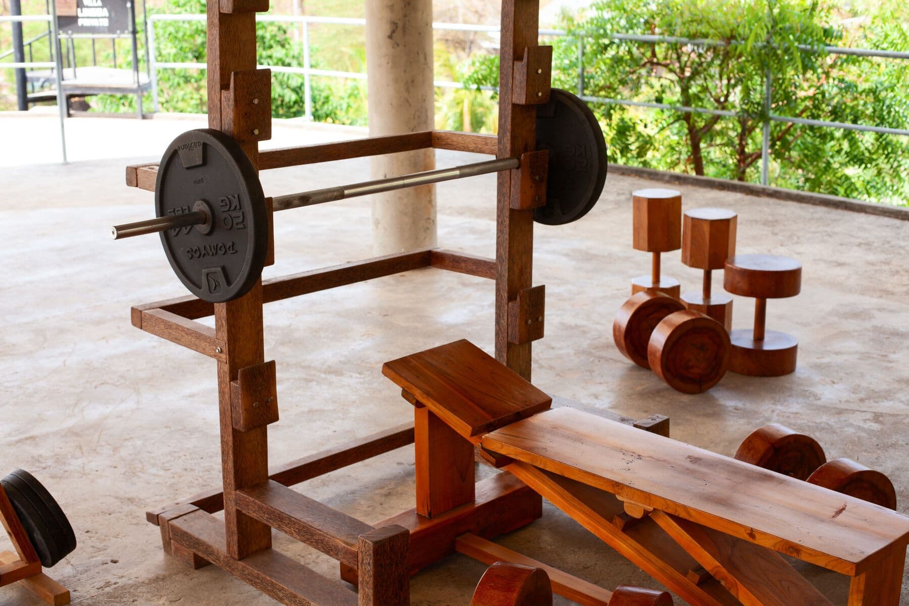 An outdoor gym with a wooden squat rack, a barbell, a bench and wooden dumbbells on a concrete floor