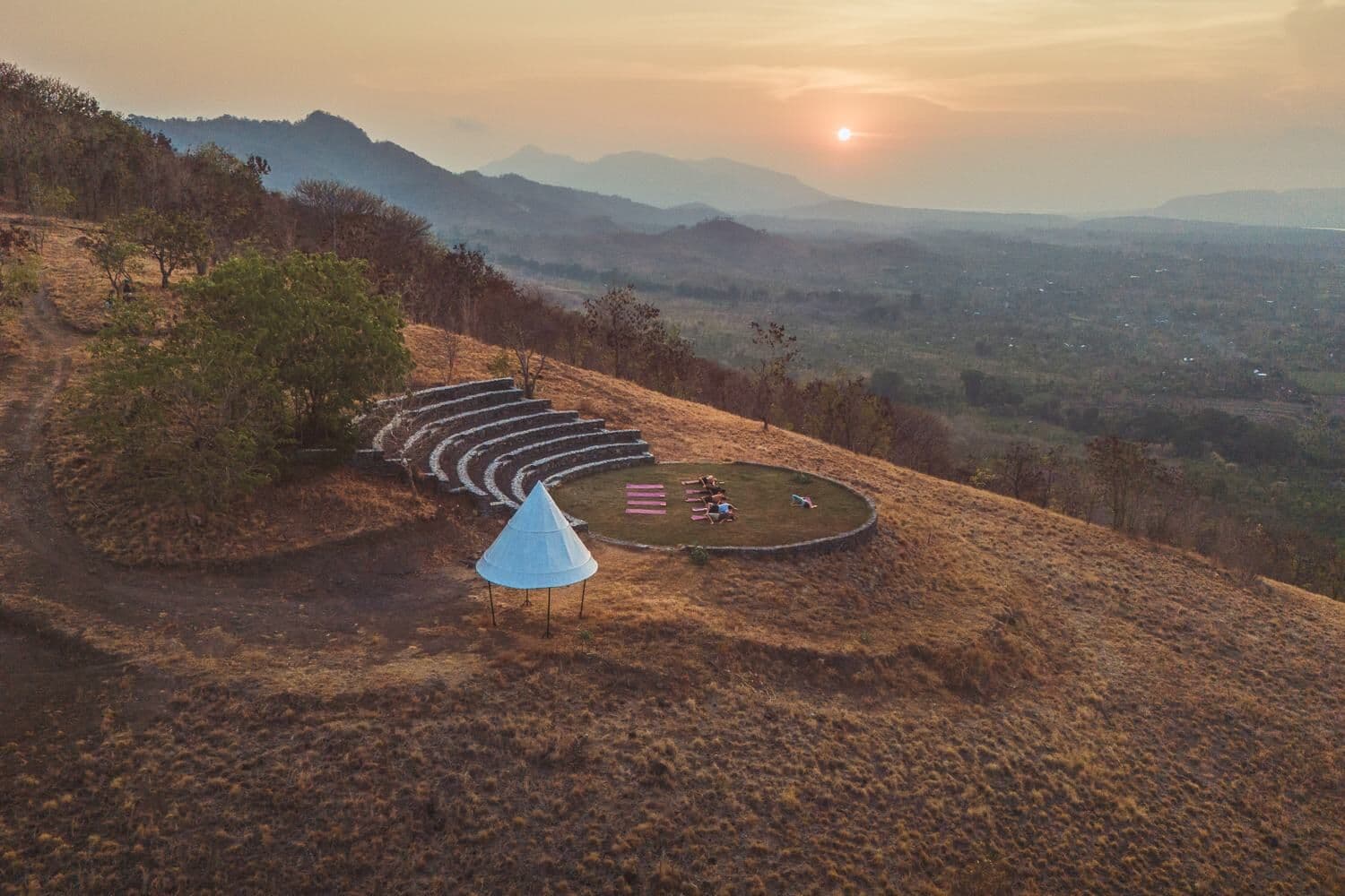 An aerial view of the hilltop amphitheatre with yoga mats laid out at sunset, mountains behind